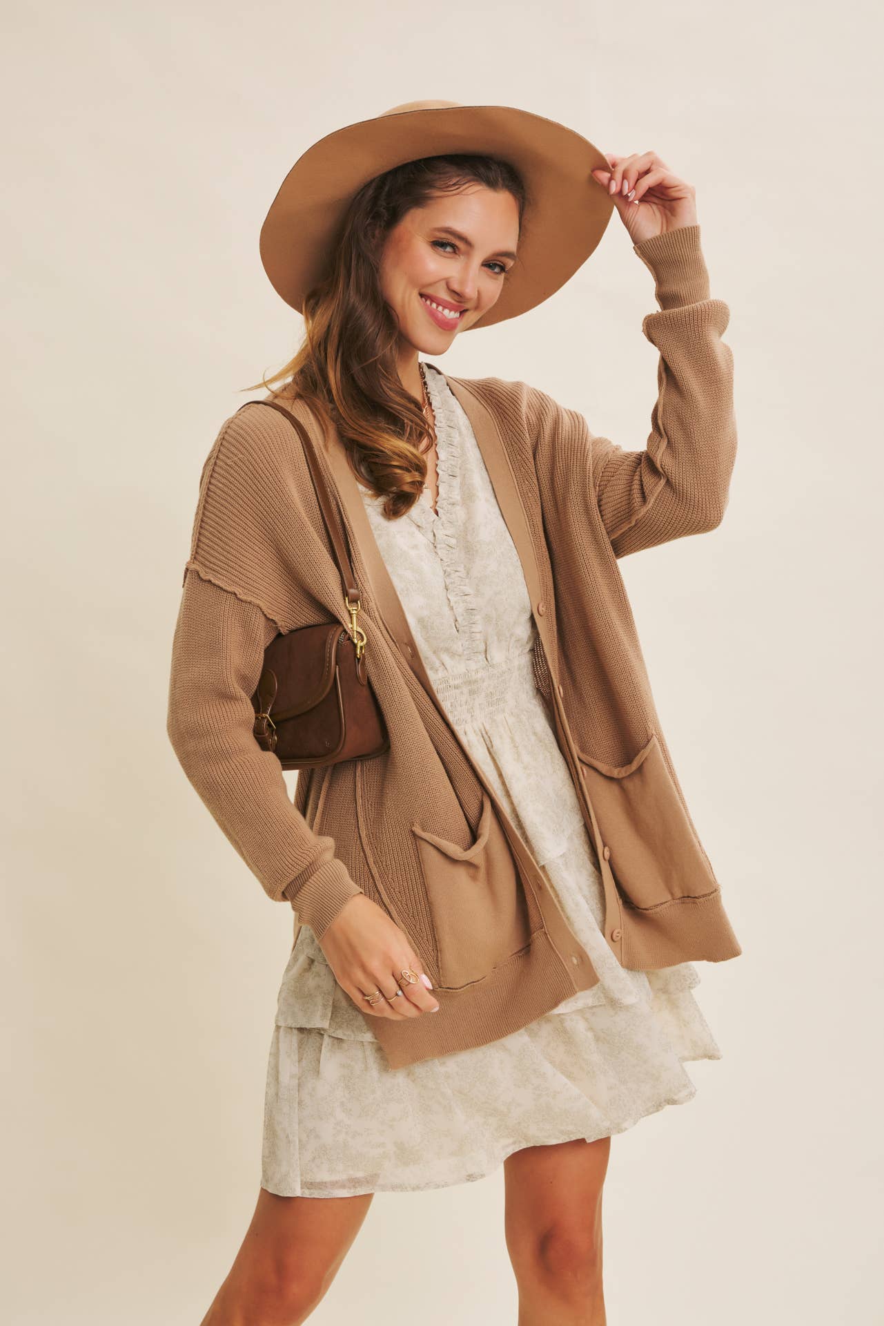 Woman wearing a beige cardigan, white dress, and brown hat against a plain background