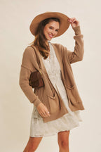 Woman wearing a beige cardigan, white dress, and brown hat against a plain background